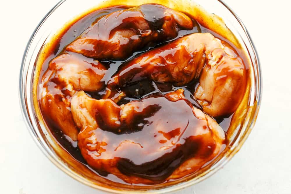 This grilled BBQ chicken is a simple to make tender chicken that's coated in a sweet homemade barbecue sauce that comes through with great flavor in every bite!ย You will be making this weekly after you see what a huge hit it is! Chicken marinating in barbecue sauce in a glass bowl.