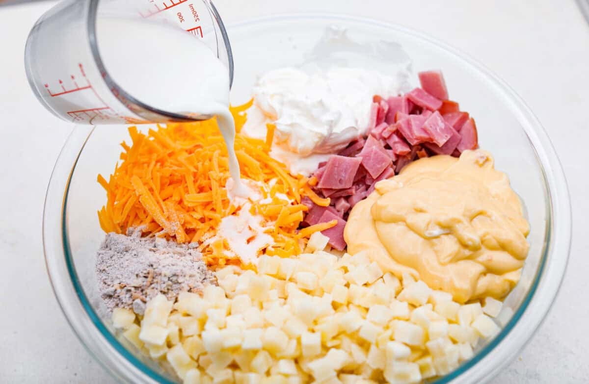 Toss your leftover ham in the slow cooker with potatoes, melty cheese, and buttery stuffing for my ridiculously easy Slow Cooker Ham and Potato Casserole! Someone pouring milk into a bowl with ham, soup, potatoes, cheese, sour cream, and seasoning mix.
