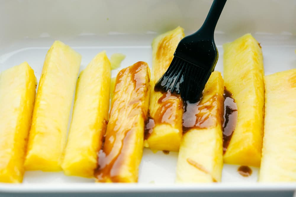 Pineapple spears that get coated in a buttery brown sugar cinnamon glaze and caramelize on the grill! Thisย will be one of the most amazing sides that you grill this summer! Pineapple spears lined up in a white dish getting brushed with a caramelized glaze.