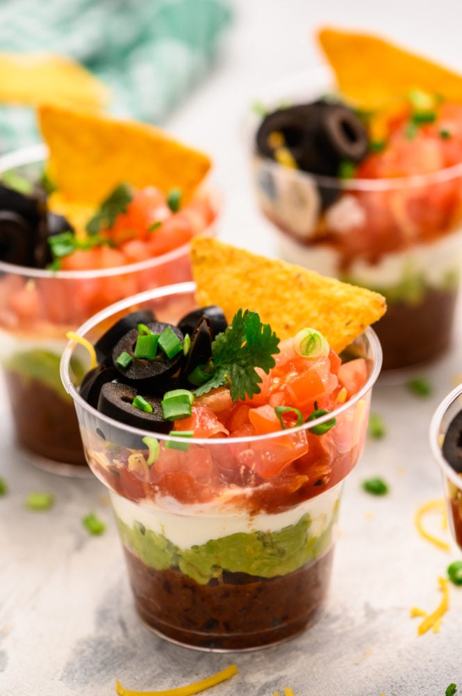 Individual Seven Layer Dip cups are perfect for layering refried beans, fresh guacamole, creamyย sourย cream, fresh salsa, chopped fresh tomatoesย and garnished with black olives, a single chip and scallions. These are ALWAYS a hit at parties! Individual cups with seven layers of refried beans, guacamole, sour cream and topped with tomatoes, black olives, scallions and a chip.