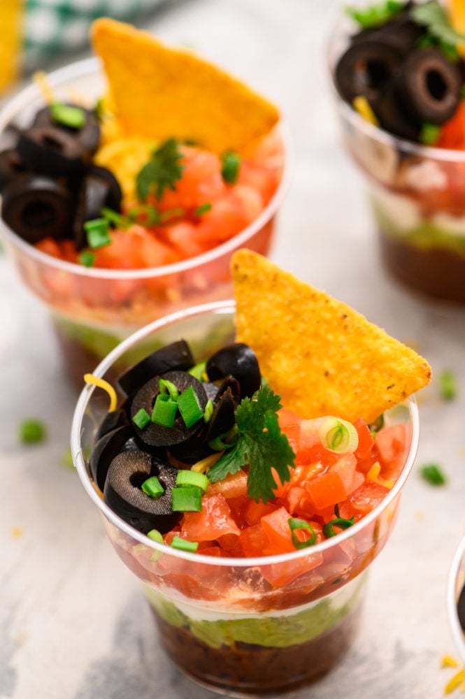 Individual Seven Layer Dip cups are perfect for layering refried beans, fresh guacamole, creamyย sourย cream, fresh salsa, chopped fresh tomatoesย and garnished with black olives, a single chip and scallions. These are ALWAYS a hit at parties! Individual cups with seven layer dip inside with black olives, tomatoes, scallions and a chip on top.
