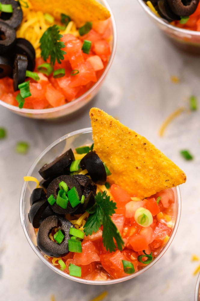 Individual Seven Layer Dip cups are perfect for layering refried beans, fresh guacamole, creamyย sourย cream, fresh salsa, chopped fresh tomatoesย and garnished with black olives, a single chip and scallions. These are ALWAYS a hit at parties! A photo of the individual cup with black olives, tomatoes, scallions and a chip.