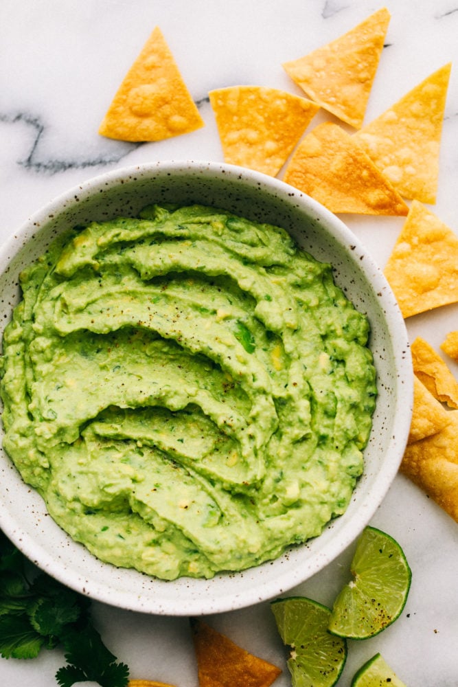 This Creamy Avocado dip is one of the best dips that I have EVER had! ย Creamy, healthy, and full of flavor, you won't be able to get enough! creamy avocado dip in a bowl with chips and lime slices around the edges.
