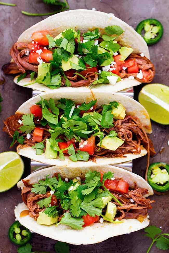 Flank steak that is rubbed in spices and slow cooks with peppers, jalapeรฑo, and onions all day to tender and juicy perfection! This recipe is easy and will become a family favorite!ย Slow cooker beef tacos with cilantro garnished on top with limes on the side.