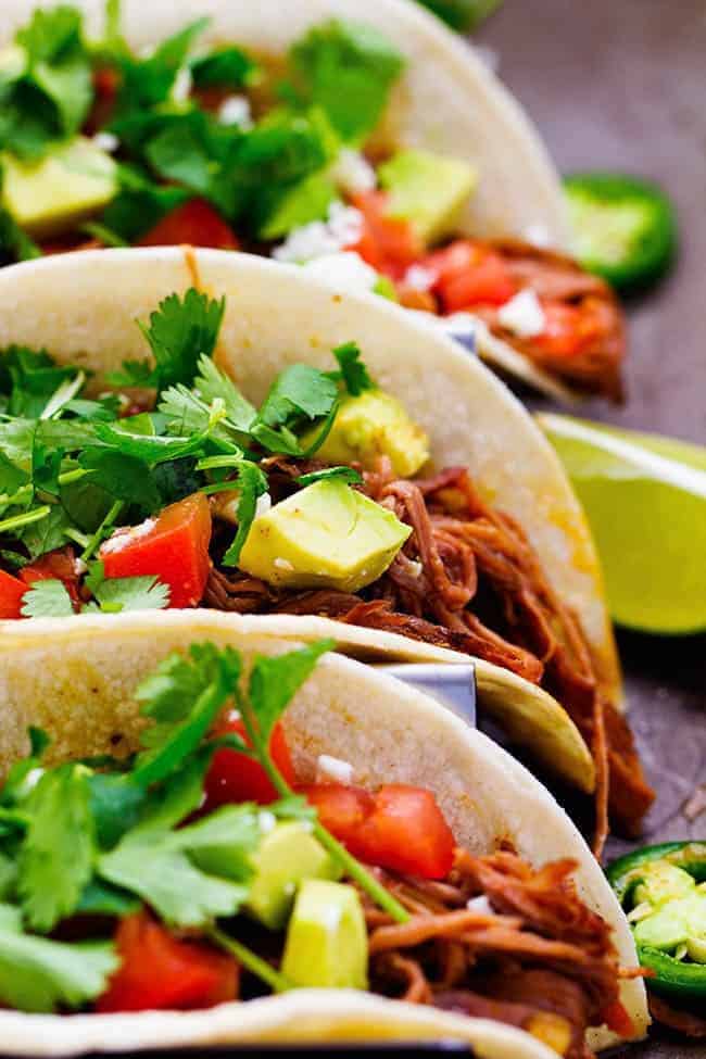 Flank steak that is rubbed in spices and slow cooks with peppers, jalapeรฑo, and onions all day to tender and juicy perfection! This recipe is easy and will become a family favorite!ย Slow cooker beef carnitas with cilantro on top and limes on the side.