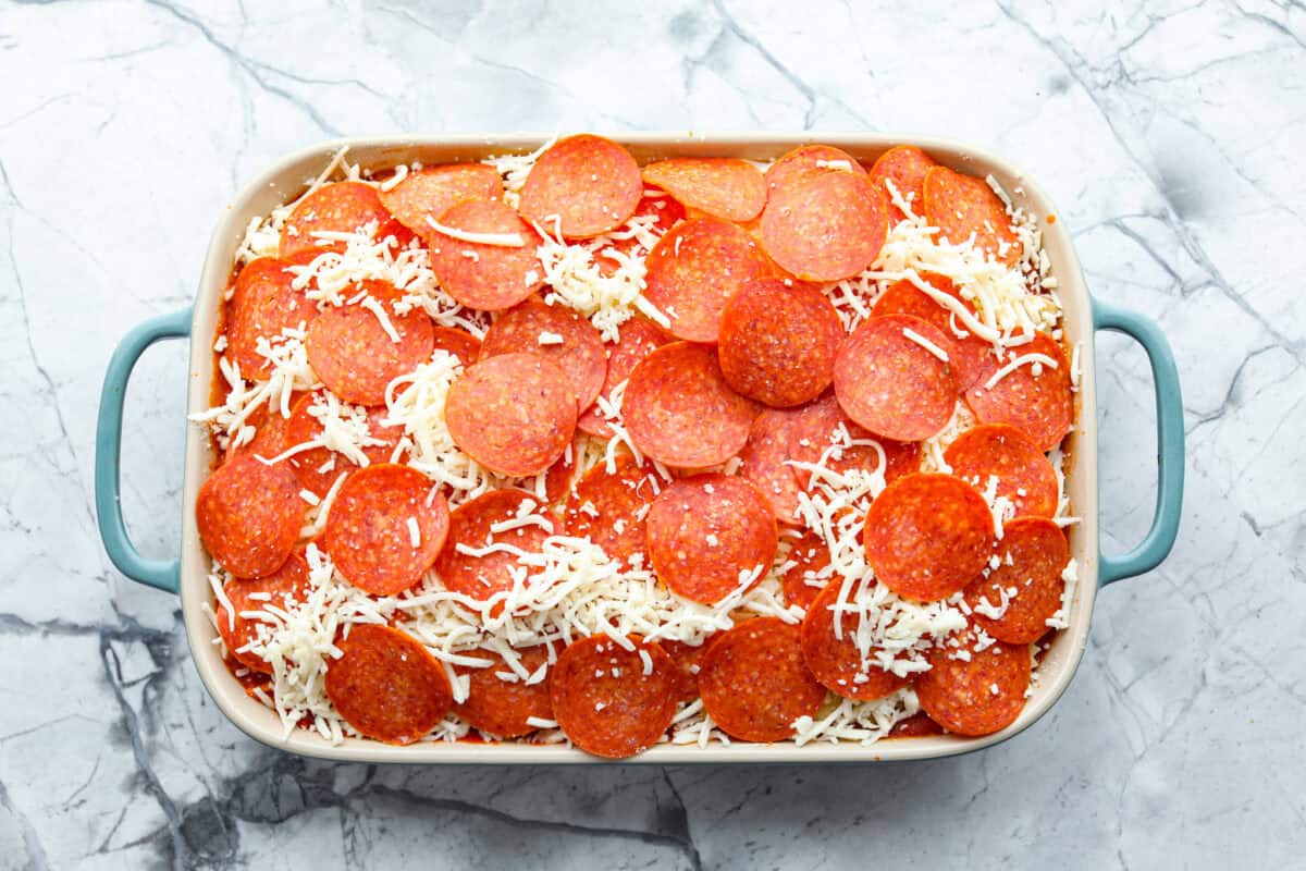 Cheesy pizza casserole combines rich tomato sauce, savory garlic, melted cheese, ham, sausage, and pepperoni. It has all the classic pizza flavors you love in a comforting casserole. Itโs a hearty, satisfying meal thatโs sure to hit the spot! Overhead shot of baking dish with uncooked pizza casserole ready to go in the oven.