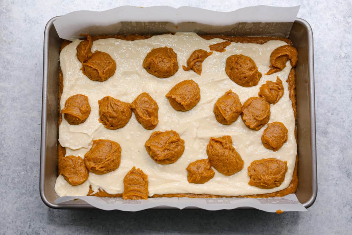 Pumpkin roll bars have all the things you love about pumpkin rolls without all of the hassle! They have perfectly spiced pumpkin cake with a smooth cream cheese swirl that will be sure to make this your new favorite fall dessert! Overhead shot of baking pan with layer of pumpkin roll mixture, topped with cream cheese mixture with dollops of more pumpkin roll mixture on top.
