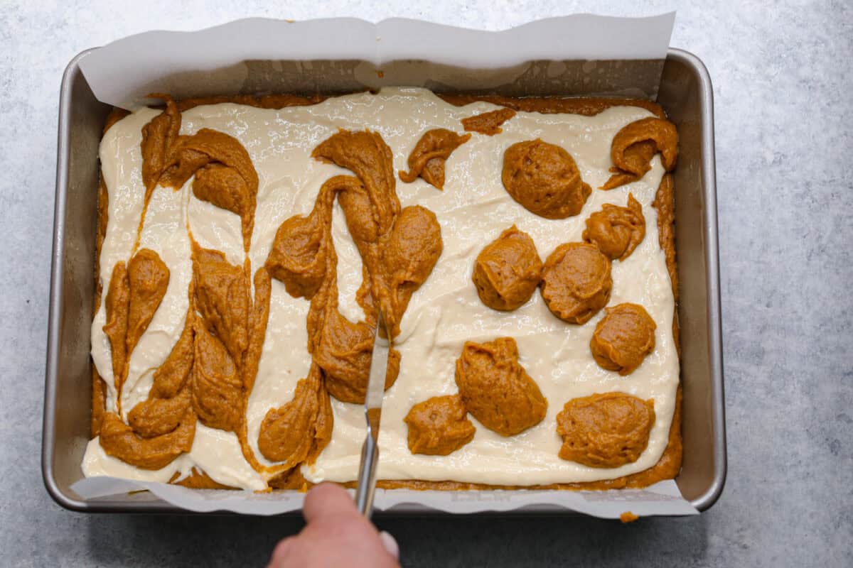 Pumpkin roll bars have all the things you love about pumpkin rolls without all of the hassle! They have perfectly spiced pumpkin cake with a smooth cream cheese swirl that will be sure to make this your new favorite fall dessert! Overhead shot of someone swirling the dollops of pumpkin roll into the cream cheese layer.