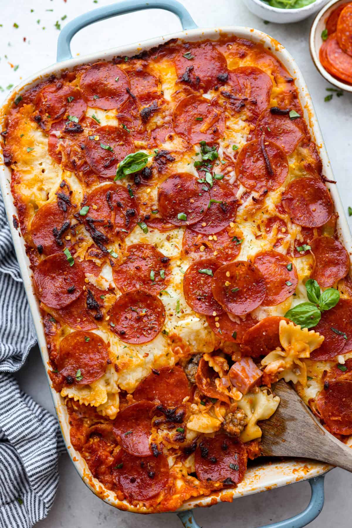Cheesy pizza casserole combines rich tomato sauce, savory garlic, melted cheese, ham, sausage, and pepperoni. It has all the classic pizza flavors you love in a comforting casserole. Itโs a hearty, satisfying meal thatโs sure to hit the spot! Overhead shot of cooked pizza casserole with wooden spoon inserted into the dish.