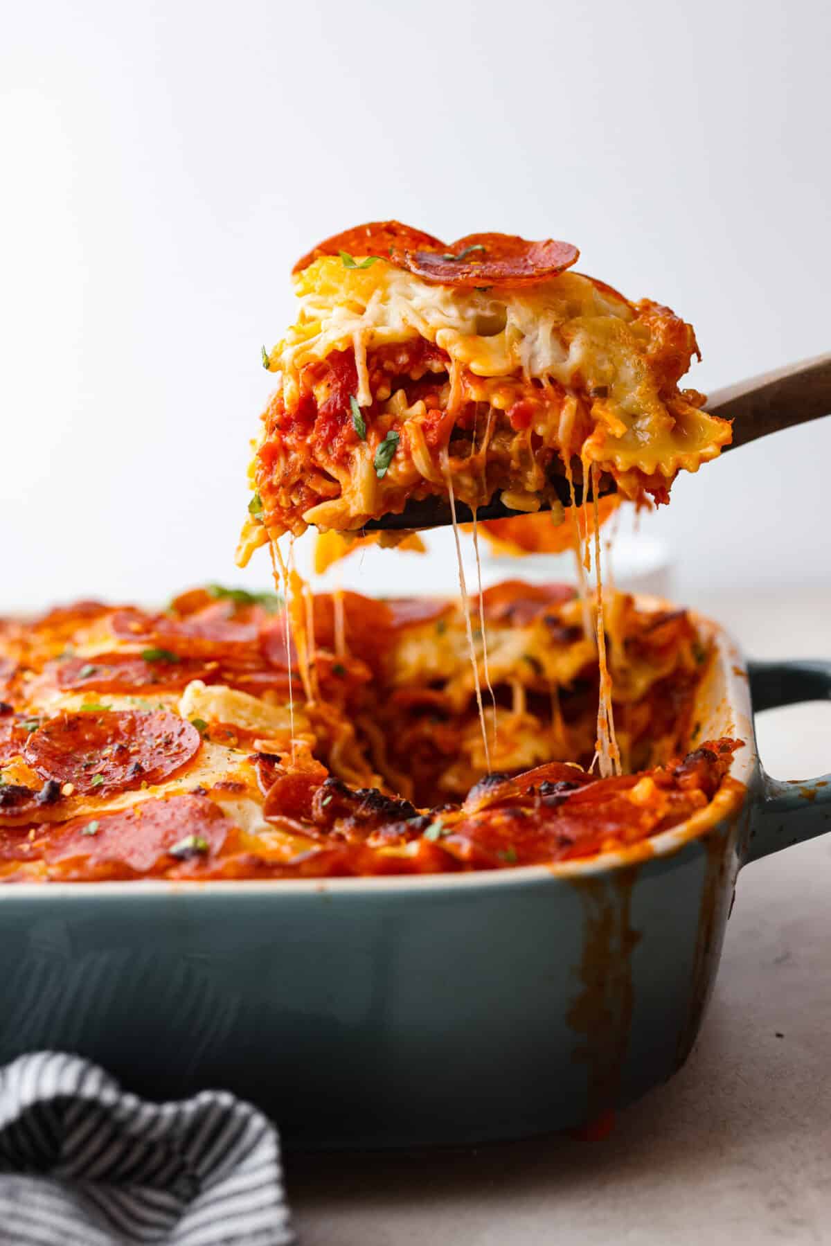 Cheesy pizza casserole combines rich tomato sauce, savory garlic, melted cheese, ham, sausage, and pepperoni. It has all the classic pizza flavors you love in a comforting casserole. Itโs a hearty, satisfying meal thatโs sure to hit the spot! Side shot of a slice of pizza casserole being lifted out of the pan with cheesy stringiness.