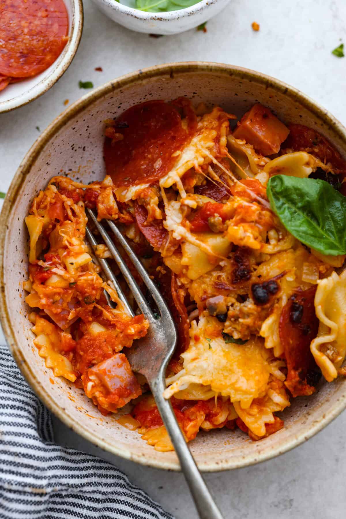 Cheesy pizza casserole combines rich tomato sauce, savory garlic, melted cheese, ham, sausage, and pepperoni. It has all the classic pizza flavors you love in a comforting casserole. Itโs a hearty, satisfying meal thatโs sure to hit the spot! Overhead shot of a bowl with a serving of cheesy pizza casserole in it with a fork and basil leaf.