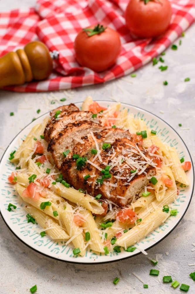 Cajun chicken pasta is so creamy and rich with a spicy kick that keeps you digging in for more.ย It is a great way to mix up your usual weekly meals.ย The cajun spice will wake up your tastebuds and your night! It is a meal you will be excited to make and even more excited to eat! Cajun chicken pasta on a white plate with two tomatoes in the background.