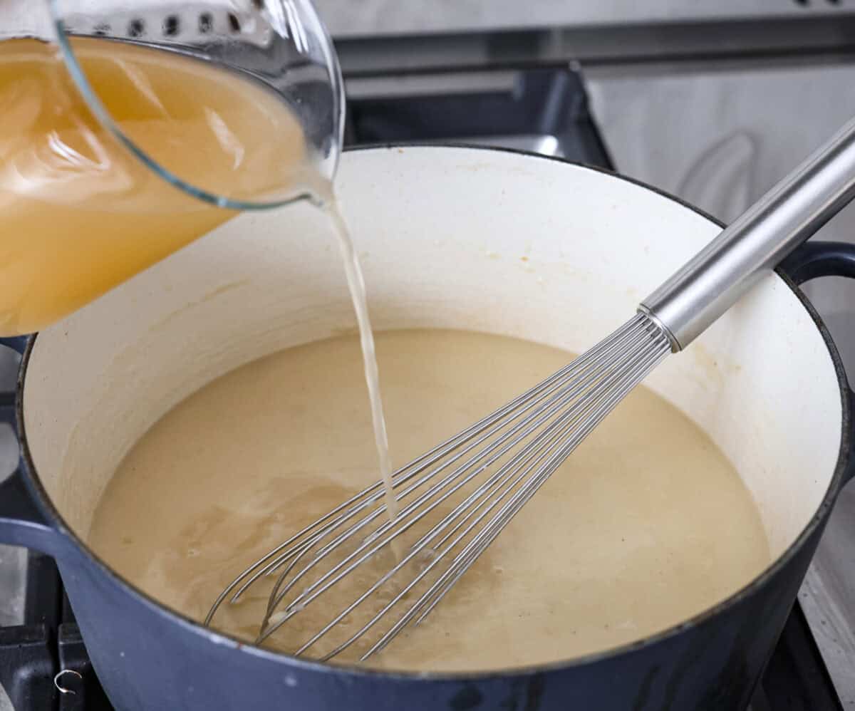 Creamy Potato Soup is the ultimate comfort food. It has a velvety, creamy base with blended potato, tender potato bites, onions, and garlic, all topped with crispy bacon! It's the perfect meal to cozy up with on a cold day. Angle shot of someone pouring in chicken broth into the roux.