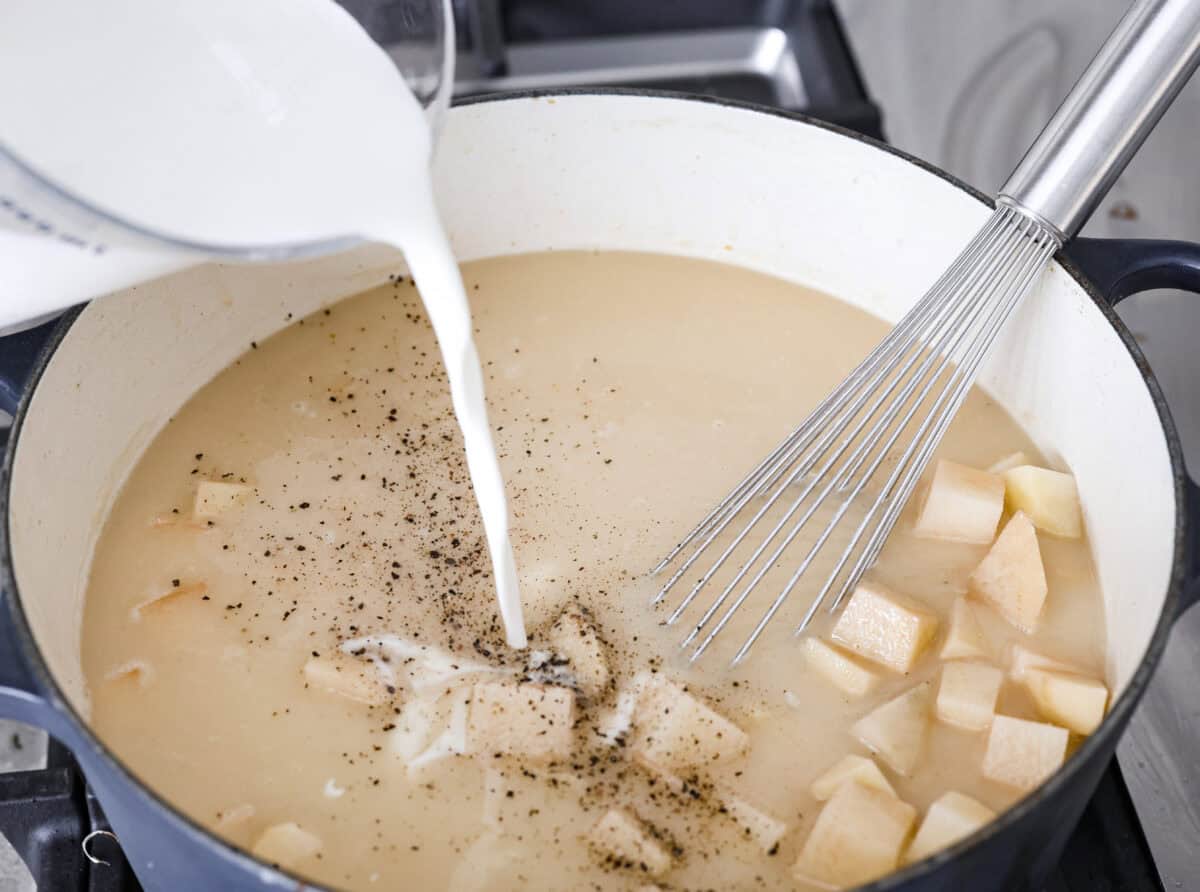 Creamy Potato Soup is the ultimate comfort food. It has a velvety, creamy base with blended potato, tender potato bites, onions, and garlic, all topped with crispy bacon! It's the perfect meal to cozy up with on a cold day. Angle shot of someone pouring the half and half into the soup mixture.