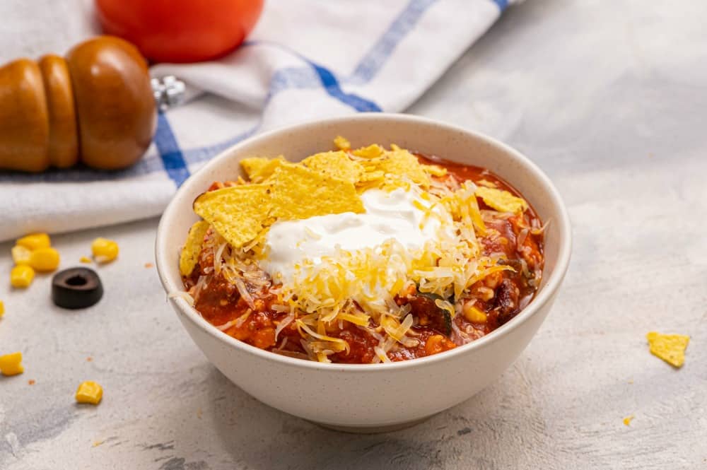 Skinny Taco Soup is jam packed with flavor and texture and is a comforting hearty meal your whole family will love! The meat and beans are super filling and the spices from the seasonings make your taste buds smile! Its the perfect complete meal all in one bowl! Skinny taco soup in a white bowl garnished with tortilla chips and shredded cheese.
