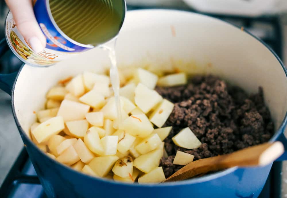 Award Winning Cheeseburger Soup is a thick and hearty soup with lean, tender ground beef, shredded carrots,ย dicedย celery and potatoes in a warm and creamy cheese soup. This is an award winning soup and it is excellent!ย Potatoes, ground beef and vegetable in a pot with chicken broth being poured over it.