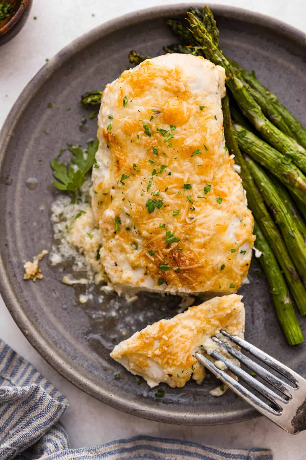 Parmesan baked chicken is so tender and juicy on the inside with a wonderful cheesy exterior. Your family will love the intense flavors, and you will love how quickly and easily this comes together! With only six ingredients and 10 minutes of prep, dinner will be served in no time! Overhead shot of plated parmesan chicken next to grilled asparagus.