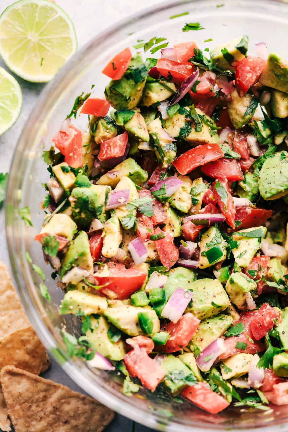 California Avocado dip is so easy to make and has so many delicious and fresh ingredients! Avocados, tomatoes, red onion, jalapeno, and cilantro come together in this irresistible dip! A zoomed in photo of the california avocado dip in a clear glass bowl with lime and tortilla chips sitting beside the bowl.