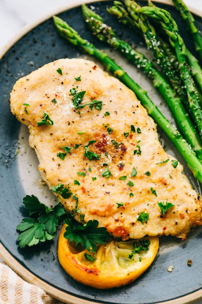 This parmesan garlic chicken is perfectly baked tender and juicy chicken that is lightly seasoned with Italian seasoning and garlic powder and then sprinkled with parmesan. All made in under 30 minutes! Baked Parmesan garlic chicken with asparagus on a plate garnished with a lemon and parsley.