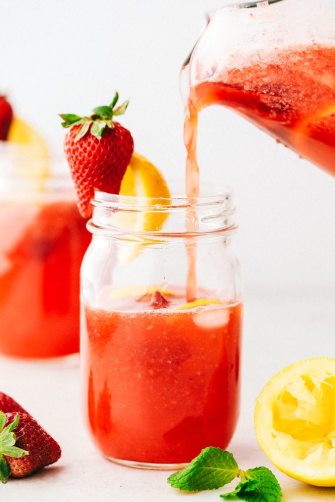 Strawberry lemonade is the perfect summertime drink that is refreshing and delicious! With only a few ingredients, this homemade lemonade will be made quickly and easily! A mason jar with strawberry lemonade being poured into it and a strawberry with a lemon stuck to the side.
