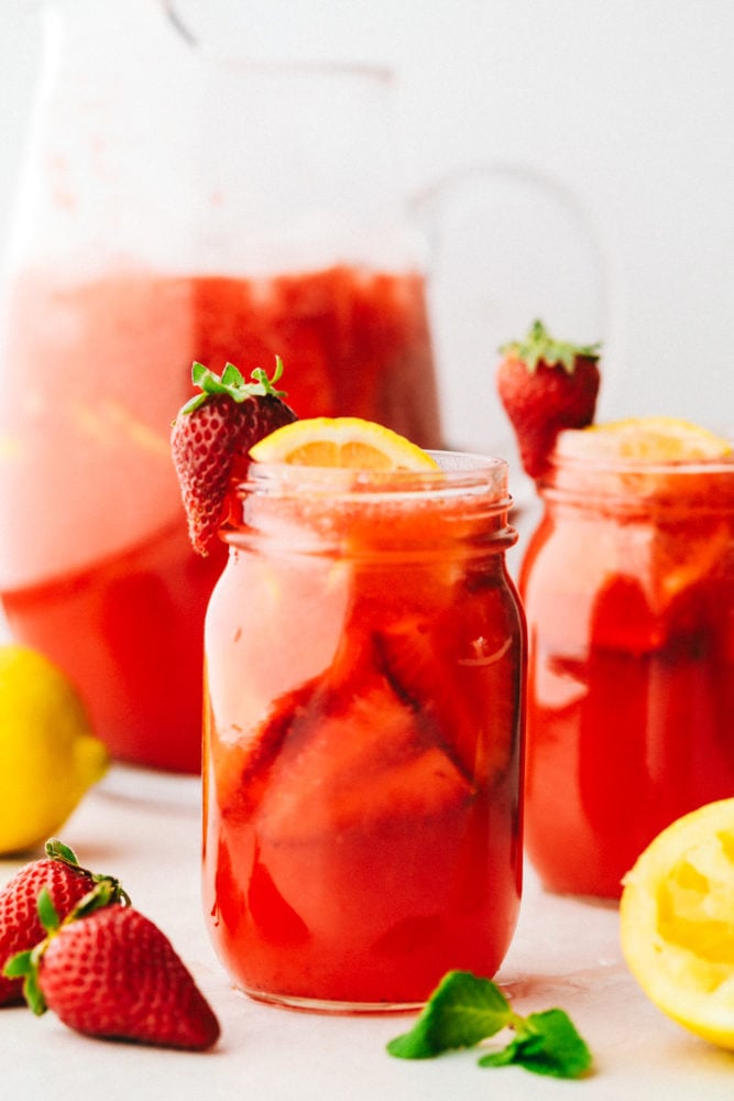 Strawberry lemonade is the perfect summertime drink that is refreshing and delicious! With only a few ingredients, this homemade lemonade will be made quickly and easily! Homemade strawberry lemonade ina a mason jar garnished with lemons and strawberries with a pitcher of lemonade in the background faded out and another mason jar with strawberry lemonade.