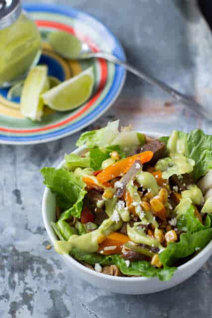 One of our favorite dinners that takes less than 30 minutes. Cheesy tortilla, fajita rice, sauteed peppers, onions and corn and avocado dressing Steak Fajita salad in a white bowl beside a plate of lime slices.