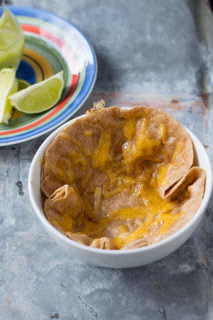 One of our favorite dinners that takes less than 30 minutes. Cheesy tortilla, fajita rice, sauteed peppers, onions and corn and avocado dressing a white bowl with a tortilla in it covered in melted cheese. A colorful plate with lime slices sits beside the bowl on the gray tray.