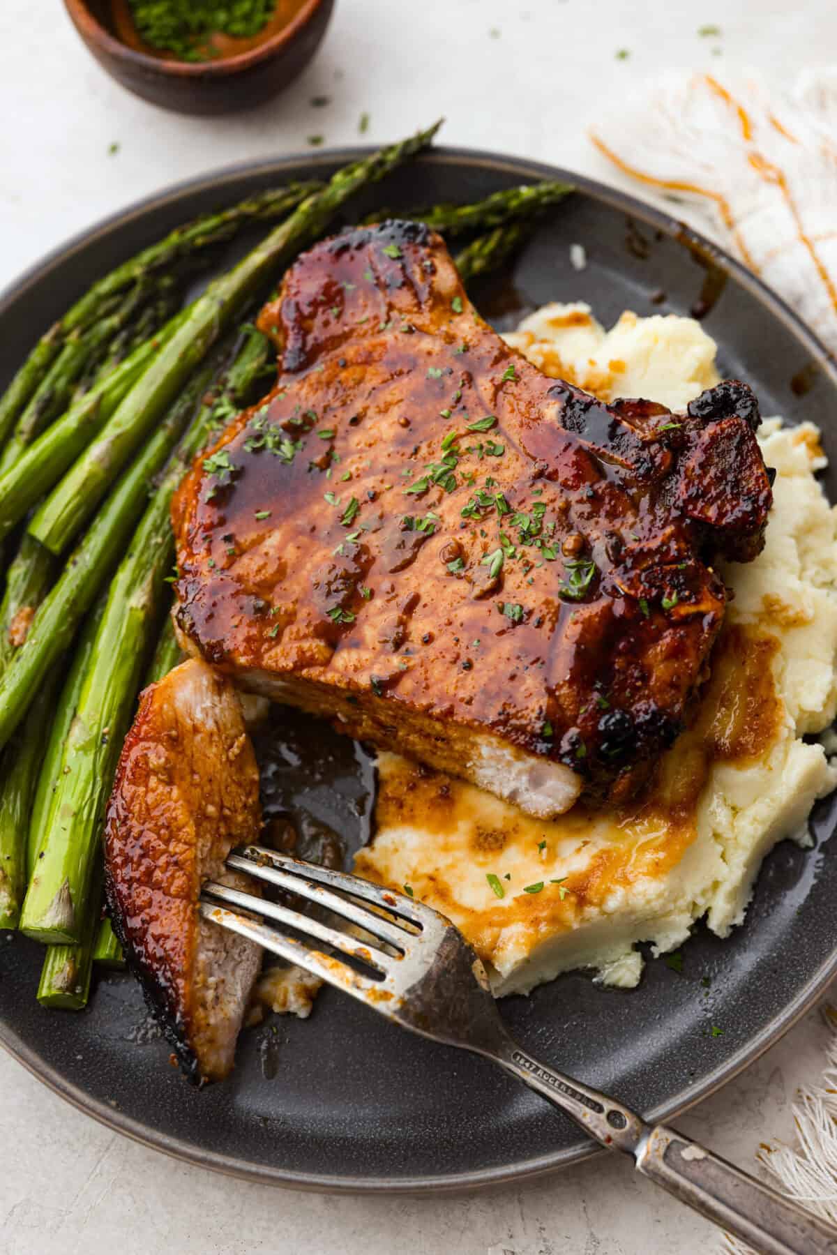 These brown sugar glazed pork chops are incredibly flavorful and delicious. Your family will love the salty pork smothered in a sweet brown sugar glaze. The crispy seared textured with the sweet sauce dripping down each bite will have you begging for more! Angle shot of plated pork chop over mashed potatoes, next to asparagus.