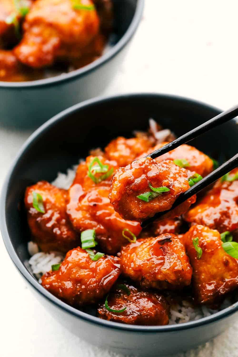 This Firecracker Chicken is sweet, spicy and adds a nice kick to your meal. The chicken is super tender and juicy and once you smother it in the flavorful sauce, you will have everyone wanting to come back for seconds! Homemade Firecracker Chicken in a black bowl over rice with some chopsticks.