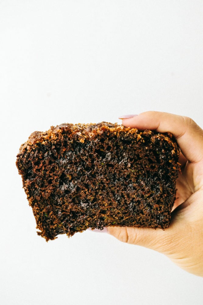 Chocolate Zucchini Bread is moist, sweet and filled with a hidden gem! It bakes perfectly and has a beautiful cracked sugar crust topping! Chocolate zucchini bread held up with a hand with a white background.