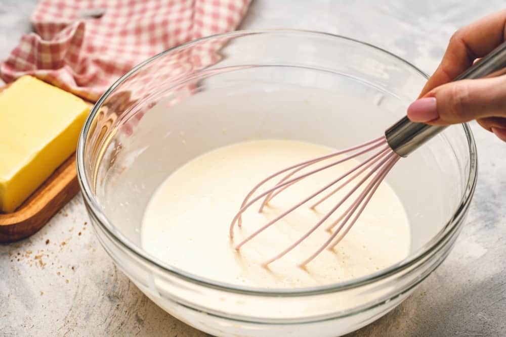 Cinnamon Roll Pancakes are aย delicious homemadeย pancake mix swirled with cinnamon roll filling and drizzled with aย delicious cream cheese glaze. These are pureย deliciousness!ย Cinnamon roll pancake mix being stirred together with a whisk in a glass bowl with a side of butter.