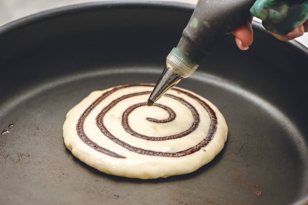 Cinnamon Roll Pancakes are aย delicious homemadeย pancake mix swirled with cinnamon roll filling and drizzled with aย delicious cream cheese glaze. These are pureย deliciousness!ย Cinnamon roll pancakes being cooked on the stove top.