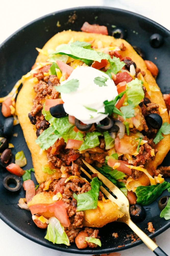 These easy and delicious Navajo tacos are made with thick, soft fry bead then layered with tasty taco fixings. A delicious family-friendly meal that you can pile high with all of your favorite toppings! Navajo tacos with ground beef, baked beans, lettuce, olives and tomatoes on top with a dollop of sour cream.