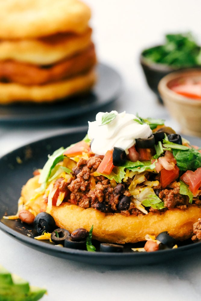 These easy and delicious Navajo tacos are made with thick, soft fry bead then layered with tasty taco fixings. A delicious family-friendly meal that you can pile high with all of your favorite toppings! Navajo taco on a black plate with beans, ground beef, tomatoes, lettuce, cheese and sour cream.
