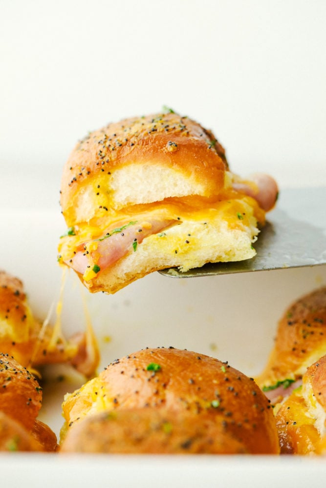 Ham and Cheddar Sliders are soft and buttery baked rolls filled with sliced ham and ooey gooey cheese. They are the perfect quick and easy crowd-pleasing meal! Pulling out a ham and cheddar slider with gooey cheese.
