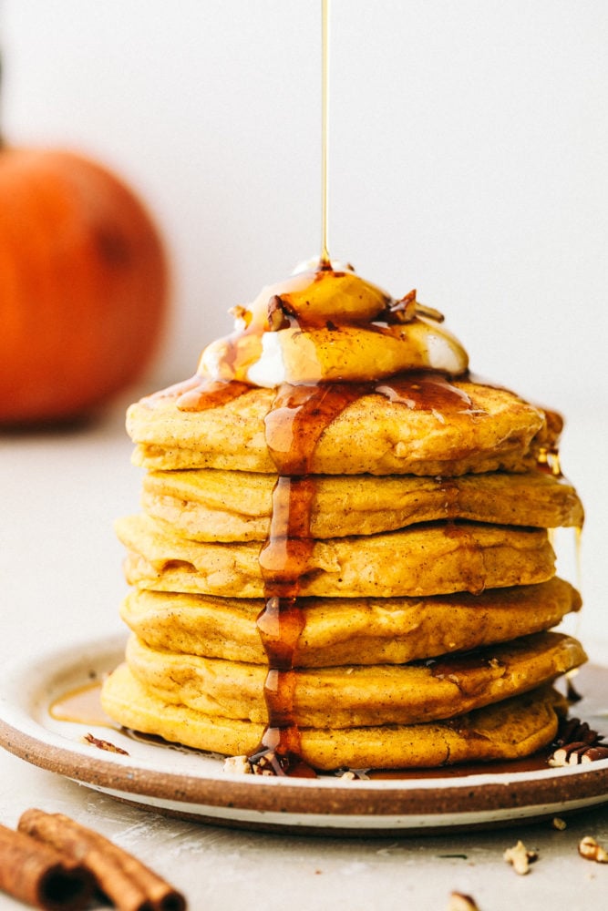 Fluffy and moist pumpkin spice pancakes are what fall breakfast dreams are made of! Perfectly spiced and full of flavor, it's like having pumpkin pie for breakfast! Side view of a stack of pumpkin spice pancakes topped with whipped cream and syrup being drizzled on top.