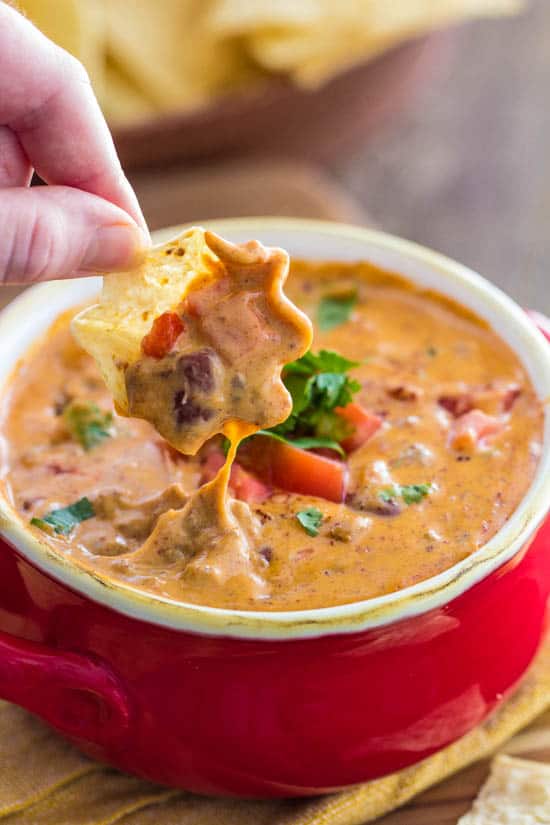 Delicious chili queso that is full of amazing flavor and is sure to be a hit! Close up of Chili Queso in a red serving cup with a person dipping a tortilla chip into it.