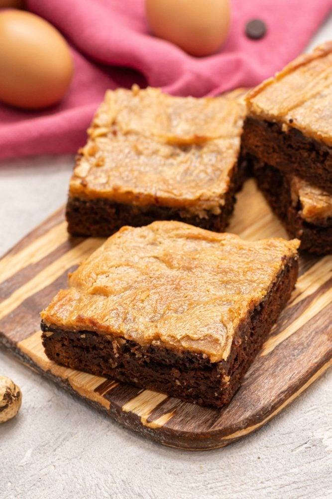 These peanut butter cheesecake brownie bars are made with moist creamy layers of dark chocolate brownie and peanut butter cheesecake.ย The yummy peanut butter and rich chocolate flavors throughout make this an irresistible treat!ย This recipe will be your first choice when you need an ooey gooey homemade dessert that everyone will go crazy over! Peanut Butter Cheesecake Brownie Bars on a wooden cutting board with two eggs in the background.