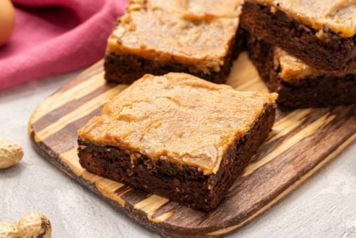 These peanut butter cheesecake brownie bars are made with moist creamy layers of dark chocolate brownie and peanut butter cheesecake.ย The yummy peanut butter and rich chocolate flavors throughout make this an irresistible treat!ย This recipe will be your first choice when you need an ooey gooey homemade dessert that everyone will go crazy over! Peanut Butter Cheesecake Brownie Bars on a wooden cutting board.