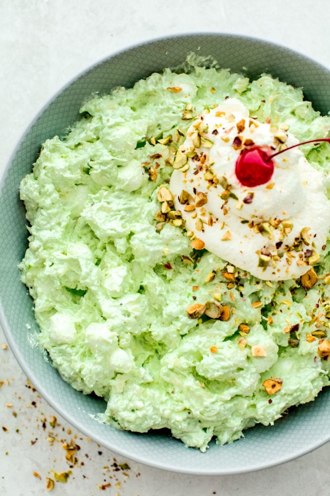 This pistachio pudding salad is a super easy and delicious dessert made with only 5 ingredients! Sweet and simple and bursting with pineapple, pistachio and marshmallow flavors, you will have a new favorite dessert salad after you try this recipe! Pistachio pudding salad in a bowl topped with whipping cream and a cherry on top.