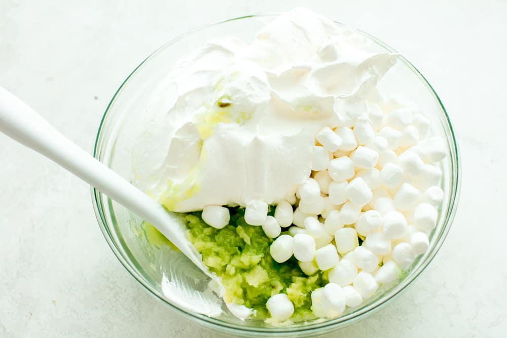 This pistachio pudding salad is a super easy and delicious dessert made with only 5 ingredients! Sweet and simple and bursting with pineapple, pistachio and marshmallow flavors, you will have a new favorite dessert salad after you try this recipe! Pistachio pudding salad ingredients in a glass bowl being stirred with a white spatula.