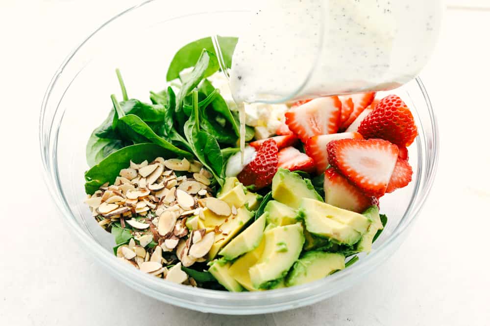 Fresh strawberries, sliced avocados, slivered almonds and sprinkled with feta cheese all tossed with a spinach salad then drizzled poppyseed dressing mixed together for the final touch. This strawberry avocado spinach salad is one everyone is raving about! Ingredients to make strawberry avocado spinach salad in a glass bowl with poppyseed dressing being poured over top.