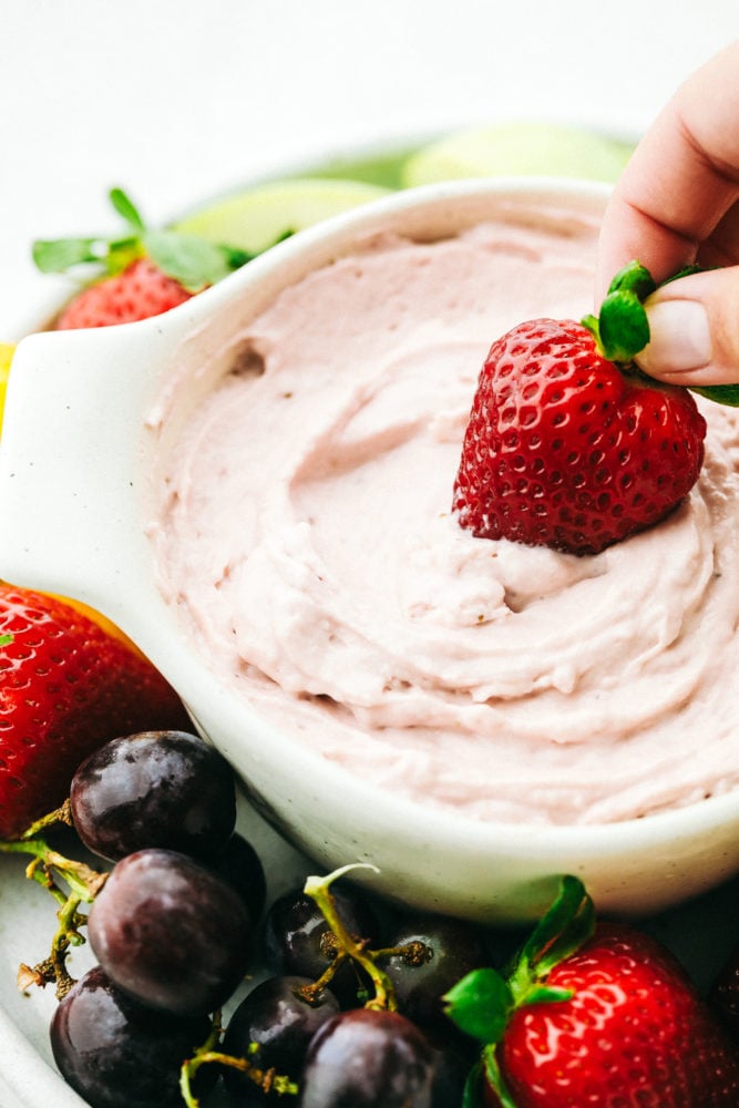 The best fruit dip that requires little effort and is absolutely fantastic! And it is only 2 ingredients! A hand dipping a large strawberry in a white dish of Strawberry Cream Cheese Fruit Dip.