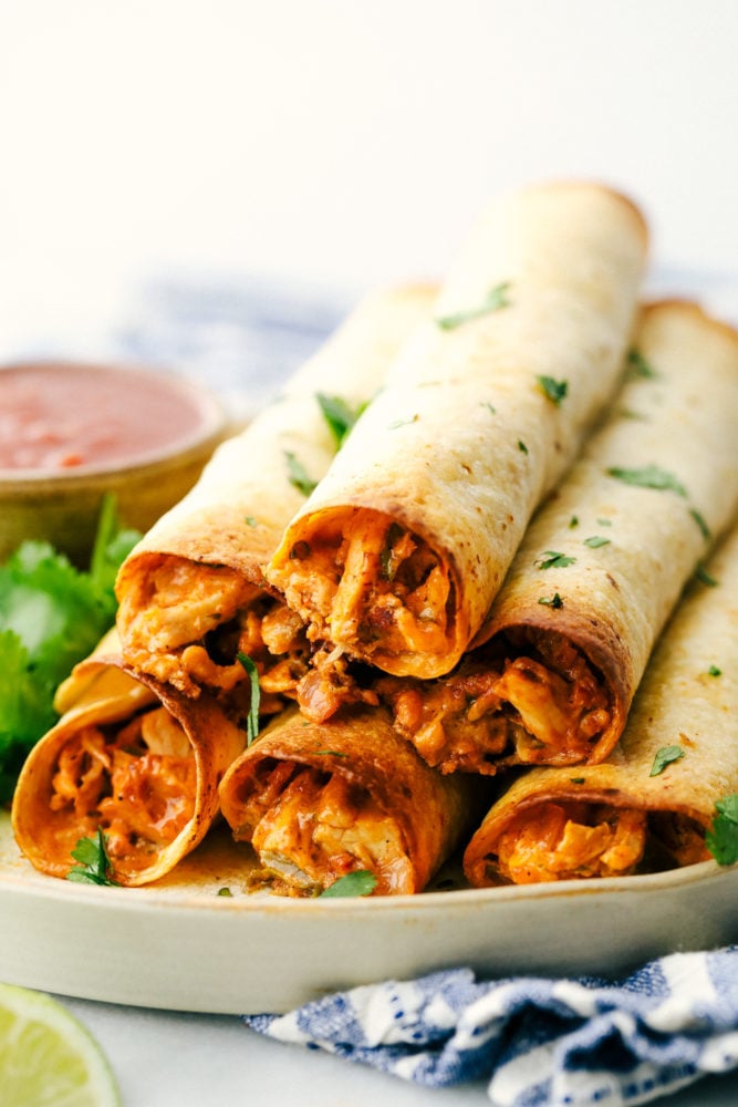 Cream cheese chicken taquitos are filled with a tender chicken mixture with cheese sprinkled over top then rolled in a flour tortilla and baked to perfection. These taquitos are a family favorite!ย Chicken taquitos stacked on top of each other on a plate.