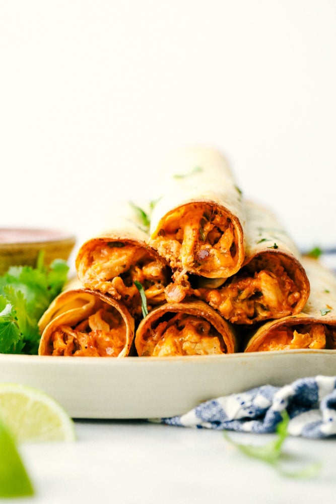 Cream cheese chicken taquitos are filled with a tender chicken mixture with cheese sprinkled over top then rolled in a flour tortilla and baked to perfection. These taquitos are a family favorite!ย Cream cheese chicken taquitos are stacked on top of each other with lime wedges as a garnish.