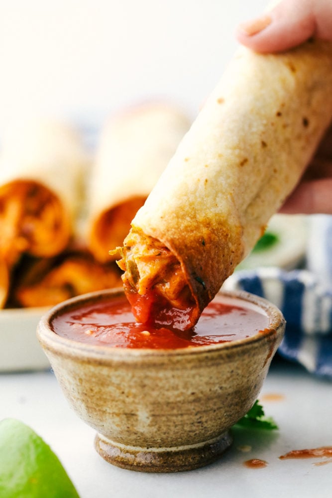 Cream cheese chicken taquitos are filled with a tender chicken mixture with cheese sprinkled over top then rolled in a flour tortilla and baked to perfection. These taquitos are a family favorite!ย Cream cheese chicken taquito is being dipped in a red salsa.