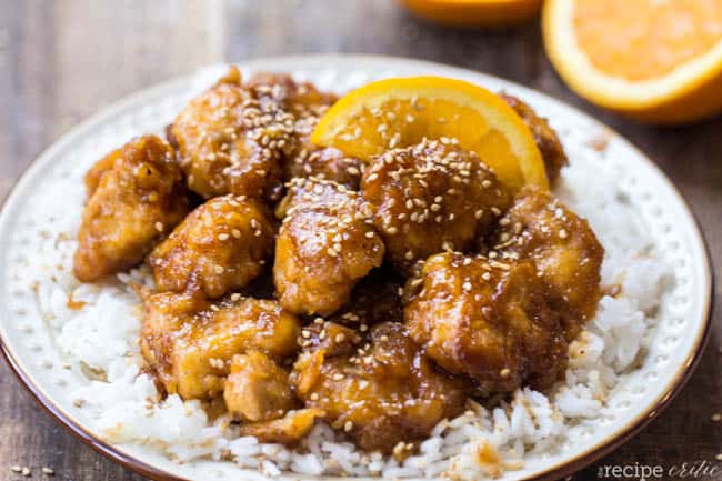 Better than take out Orange Chicken that is baked to perfection and will blow your mind! Baked orange chicken on a plate of white rice with an orange slice on the side.