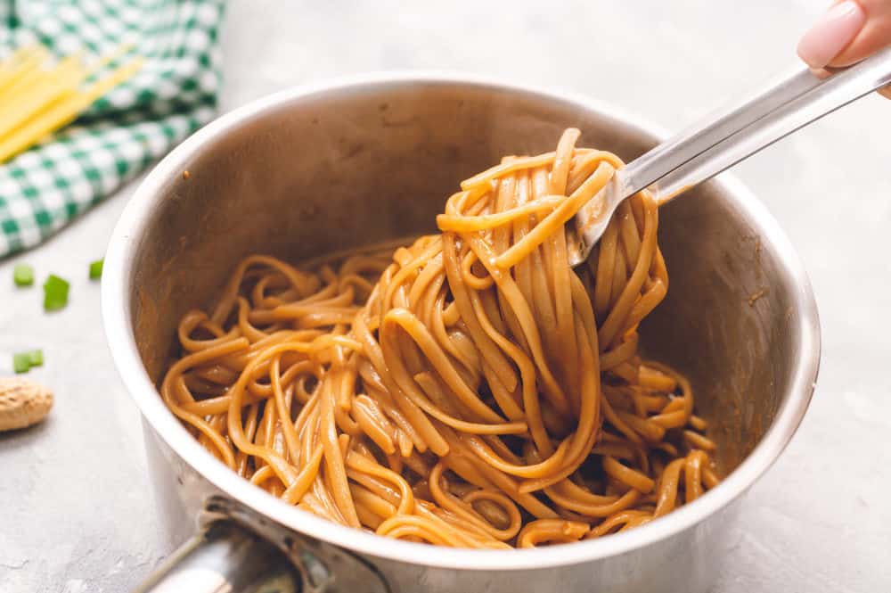 Thai Peanut Sesame Noodlesย is an asianย favorite that combines creamy peanut butter, soy sauce,ย sesame oil, minced garlic all mixed with linguine noodles tossed in this amazingly sweet and savoryย sauce.ย Thai peanut sesame noodles cooked in a pan with tongs grabbing it out of the pan.