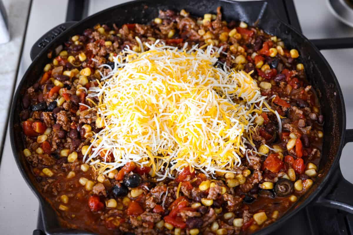 This beef enchilada dip packs all of my favorite things into one dish. It starts with red enchilada sauce, savory beef, olives, and sweet corn smothered in melty cheese. Grab your favorite tortilla chips and get dipping. Overhead shot of meet and veggie mixture with cheese on top in a skillet.