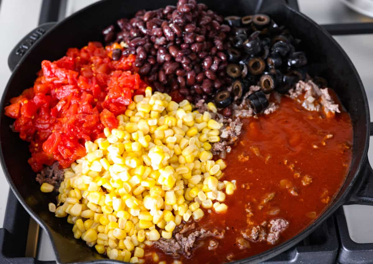This beef enchilada dip packs all of my favorite things into one dish. It starts with red enchilada sauce, savory beef, olives, and sweet corn smothered in melty cheese. Grab your favorite tortilla chips and get dipping. Overhead shot of cooked beef and onions with enchilada sauce, corn, olives, tomatoes and beans on top.